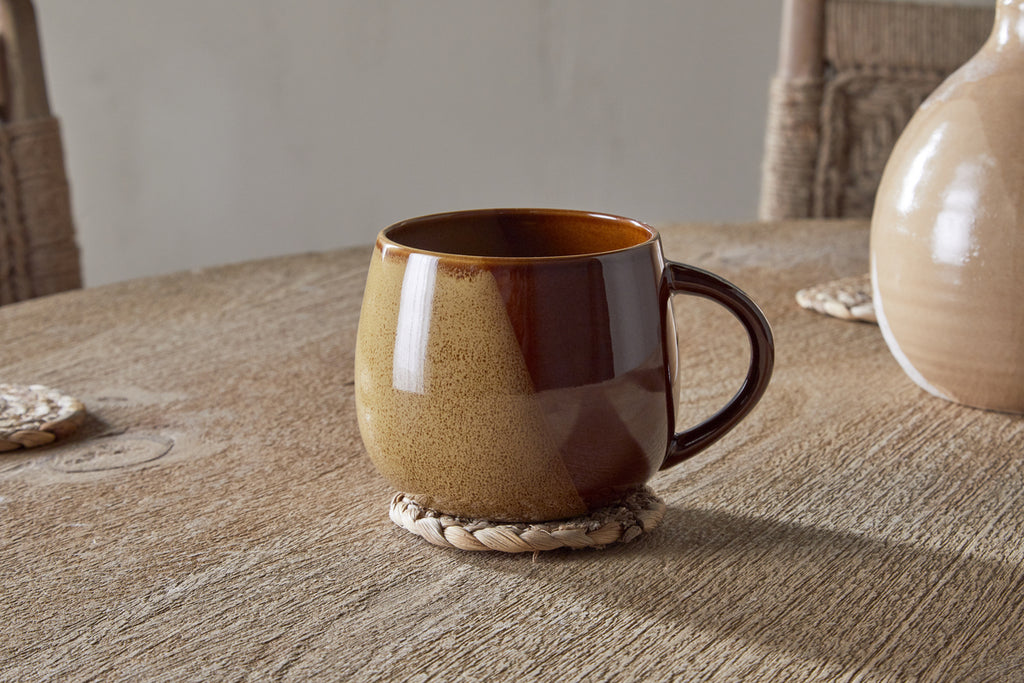 The nkuku Naav Mug - Mustard (Extra Large) features a rounded brown design, resting on a woven coaster on a wooden table, with a ceramic vase and chairs in the background.