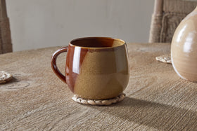 The Naav Mug - Mustard (Extra Large) by nkuku sits on a round woven coaster on a wooden table, with part of a vase and two woven placemats in the background.