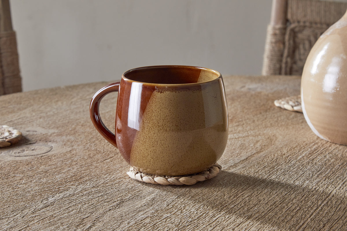 The Naav Mug - Mustard (Extra Large) by nkuku sits on a round woven coaster on a wooden table, with part of a vase and two woven placemats in the background.
