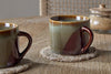 Two nkuku Naav Mugs - Blue - Small (Set of 2) are placed on woven coasters on a wooden table, with a glass jar of brown sugar and lid in the background.