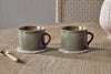 Two nkuku Naav Mugs - Blue - Large (Set of 2) with a unique glaze rest on woven coasters on a wooden table. A golden spoon with a brown handle and part of a textured vase enhance the distinctive tableware charm.