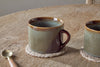 The Naav Mugs - Blue - Large (Set of 2) by nkuku rest on woven coasters atop a wooden table, accompanied by a brass spoon and part of another mug.