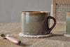The nkuku Naav Mugs - Blue - Large (Set of 2) rest on a woven coaster atop a wooden table, with a small spoon with a wooden handle nearby and part of a woven chair in the background.