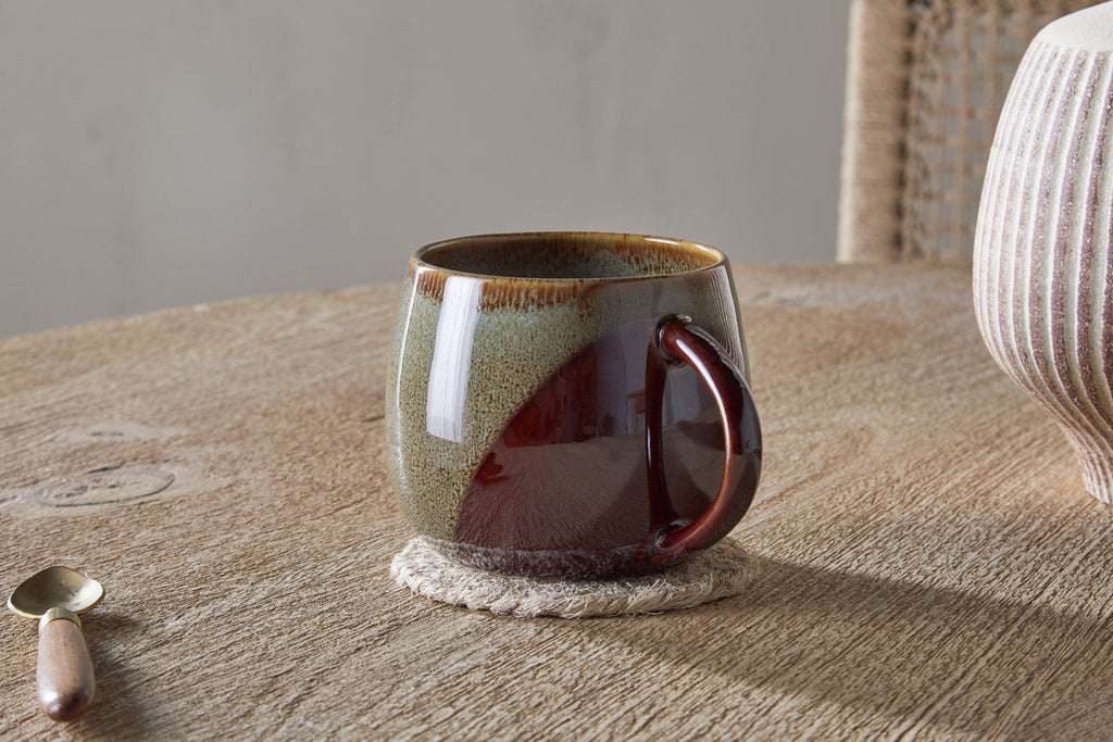 The nkuku Naav Mug - Blue (Extra Large) features a two-tone reactive glaze and sits stylishly on a coaster beside a spoon and striped vase, creating distinctive tableware for any wooden table.