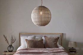 A cozy bedroom with a wooden bed, neutral bedding, and brown accent pillows features the Nilaveli Woven Jute Sphere Pendant by nkuku hanging above. A small lamp and vase with dried branches sit on a wooden nightstand against a textured off-white wall.