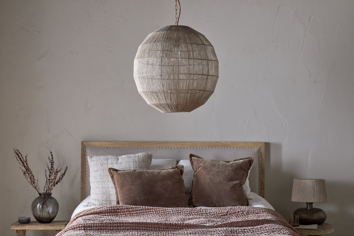 A cozy bedroom includes a wooden bed with white and brown bedding, two brown pillows, and the Nilaveli Woven Jute Sphere Pendant in Natural by nkuku hanging overhead. Wooden nightstands hold a small lamp and vase; the wall is light gray.