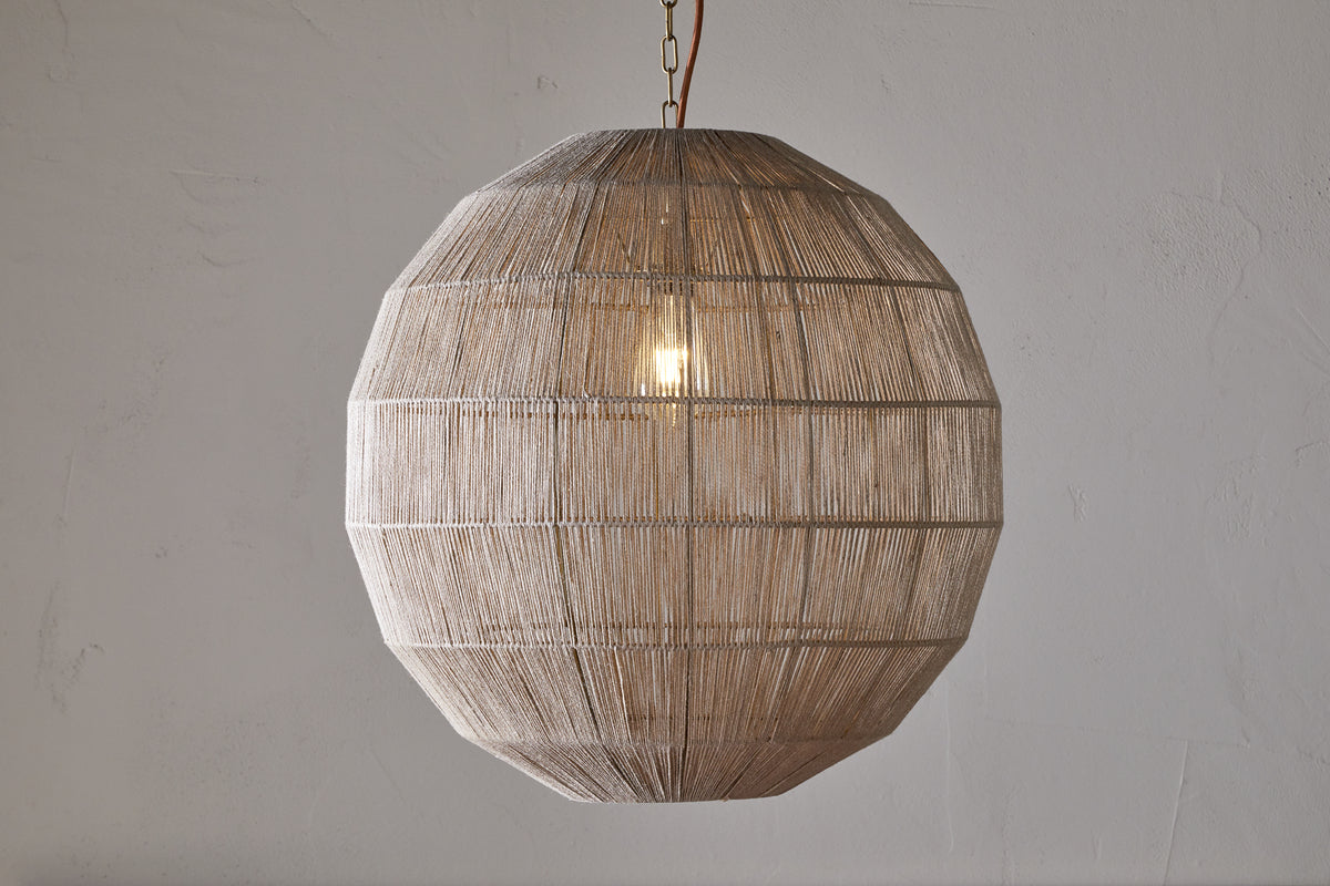 The nkuku Nilaveli Woven Jute Sphere Pendant in Natural hangs from the ceiling, its geometric, spherical jute shade showcasing a textured, see-through design that reveals the glowing bulb against a light-colored wall.