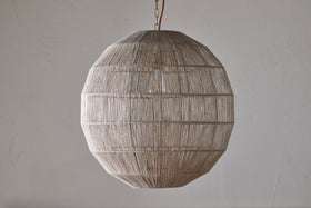 The Nilaveli Woven Jute Sphere Pendant by nkuku hangs from the ceiling, its natural jute threads tightly woven around a geometric frame. The textured, semi-transparent shade stands out against a plain, light-colored wall.