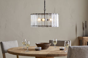 A round wooden dining table set for four with wine glasses, bowls, and a centerpiece bowl is surrounded by four upholstered chairs. Above, the nkuku Nauka Statement Pendant in aged bronze and clear hangs against a light beige textured wall.