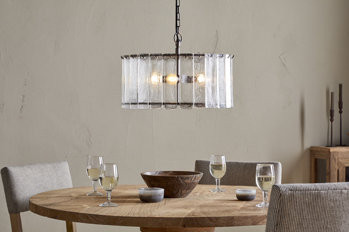 A round wooden dining table set for four with wine glasses, bowls, and a centerpiece bowl is surrounded by four upholstered chairs. Above, the nkuku Nauka Statement Pendant in aged bronze and clear hangs against a light beige textured wall.