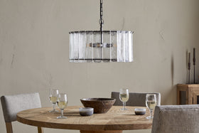 The Nauka Statement Pendant by nkuku, crafted from recycled glass with an aged bronze finish, elegantly hangs from the ceiling, making it a striking centerpiece above a table set with wine glasses.