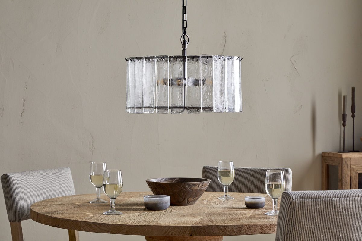 The Nauka Statement Pendant by nkuku, crafted from recycled glass with an aged bronze finish, elegantly hangs from the ceiling, making it a striking centerpiece above a table set with wine glasses.