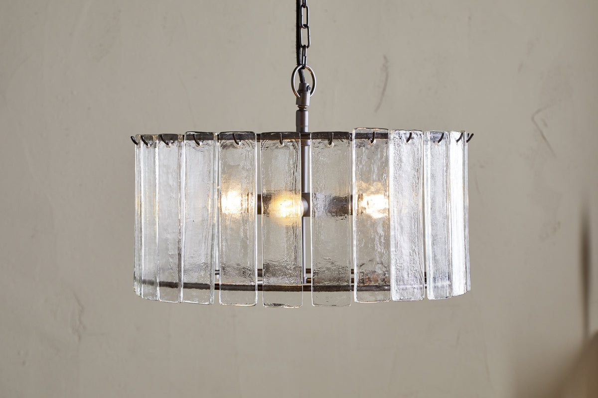The Nauka Statement Pendant from nkuku features vertical recycled glass panels forming a round shade around three bulbs. Suspended by a black chain, this aged bronze and clear chandelier creates an elegant focal point on any light wall.