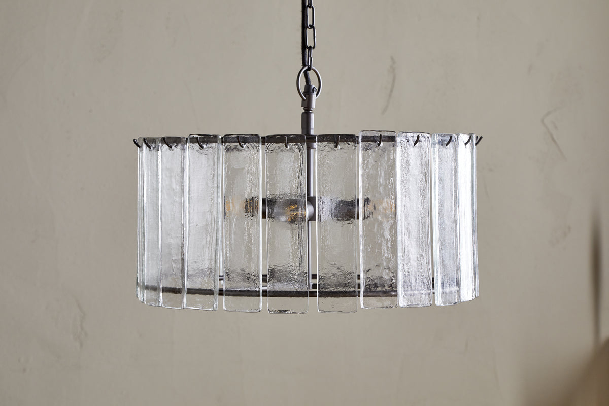 The nkuku Nauka Statement Pendant in Aged Bronze & Clear hangs from a chain, showcasing vertical textured recycled glass panels around its metal frame. Visible bulbs add elegant statement lighting against a light-colored wall.