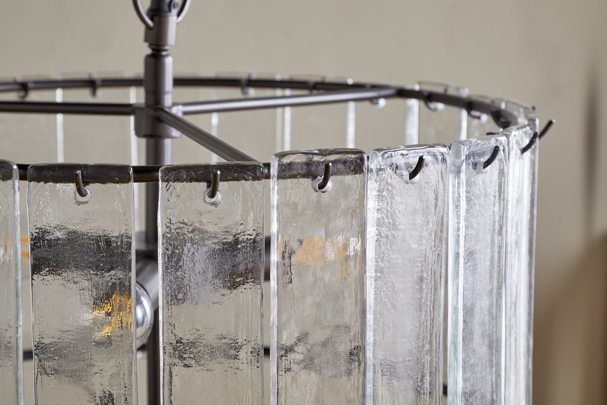 A close-up of the nkuku Nauka Statement Pendant in aged bronze and clear: vertically hanging textured recycled glass panels partly obscure glowing bulbs, creating a softly diffused light effect within a dark metal frame.
