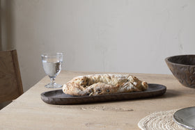 A rustic olive loaf rests on the nkuku Nyati Serving Platter - Dark Brown atop a light wood table, accompanied by a textured glass, woven placemat, wooden bowl, and chair—a minimalist scene that highlights sustainable home decor.