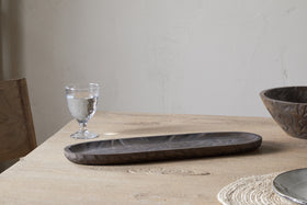 The nkuku Nyati Serving Platter in dark brown sits on a light wooden table with a clear glass and bowl, atop a woven placemat against a plain, light-colored wall.