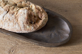 A rustic loaf sits on the Nyati Serving Platter - Dark Brown by nkuku, atop a textured wooden surface. Only part of the bread and platter are visible, adding charm to sustainable home decor.