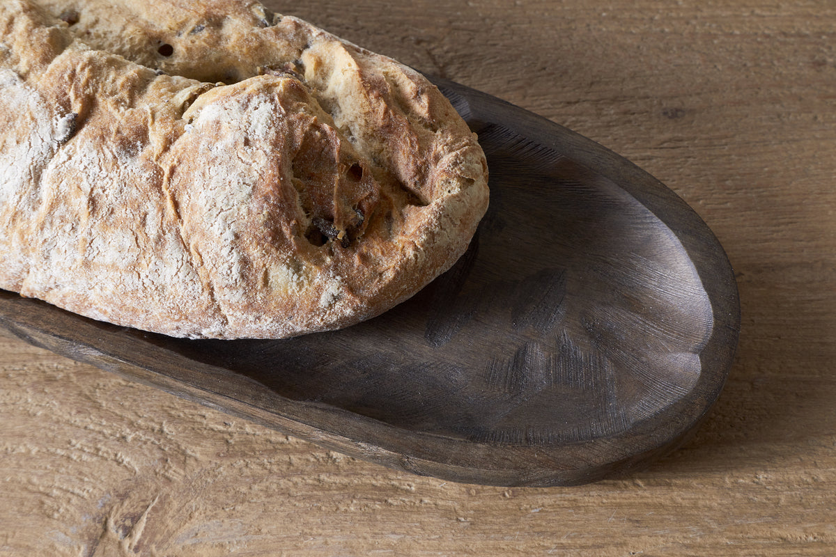 A rustic loaf sits on the Nyati Serving Platter - Dark Brown by nkuku, atop a textured wooden surface. Only part of the bread and platter are visible, adding charm to sustainable home decor.