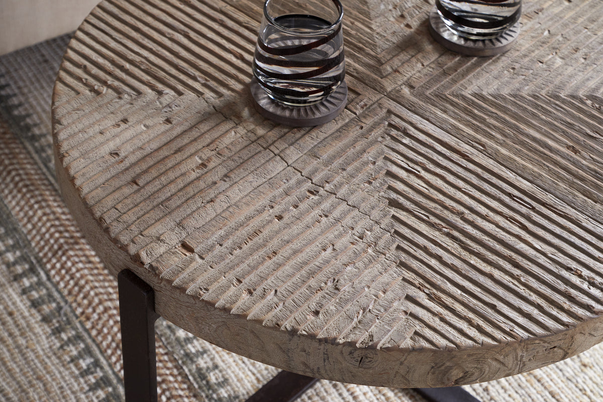 The Chakala Reclaimed Wood Coffee Table by nkuku features a textured, carved geometric top and dark iron legs. It holds two clear glasses with dark stripes and sits on a woven checkered-pattern rug partially visible below.