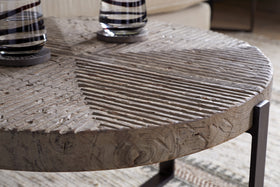A close-up of the nkuku Chakala Reclaimed Wood Coffee Table, showcasing its geometric carved top in reclaimed wood and iron legs. Two glass vases with dark stripes sit on it, all resting above a neutral textured rug.