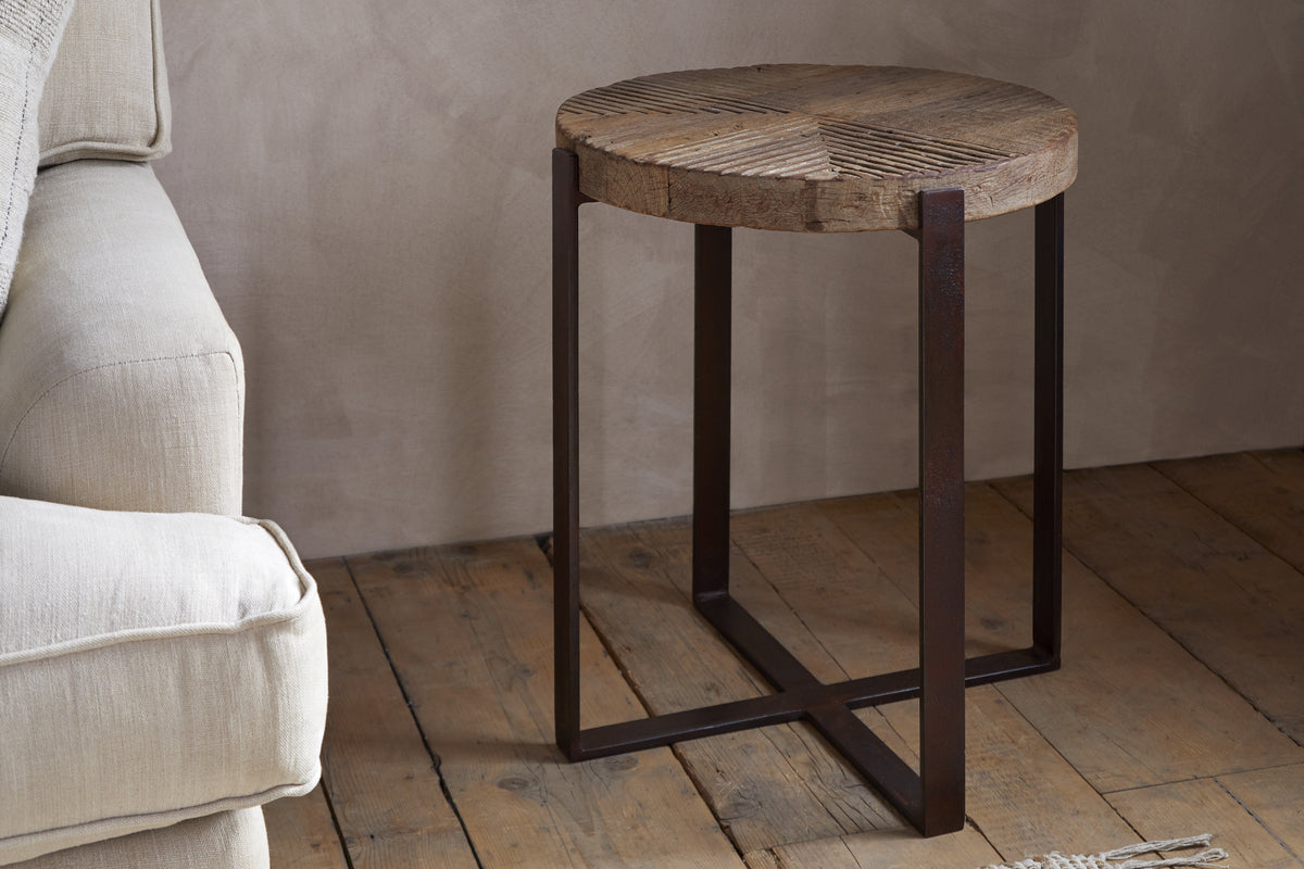 The nkuku Chakala Reclaimed Wood Side Table features a textured reclaimed wood top and dark iron legs with intersecting supports, adding modern rustic charm beside a beige sofa against a light brown wall.