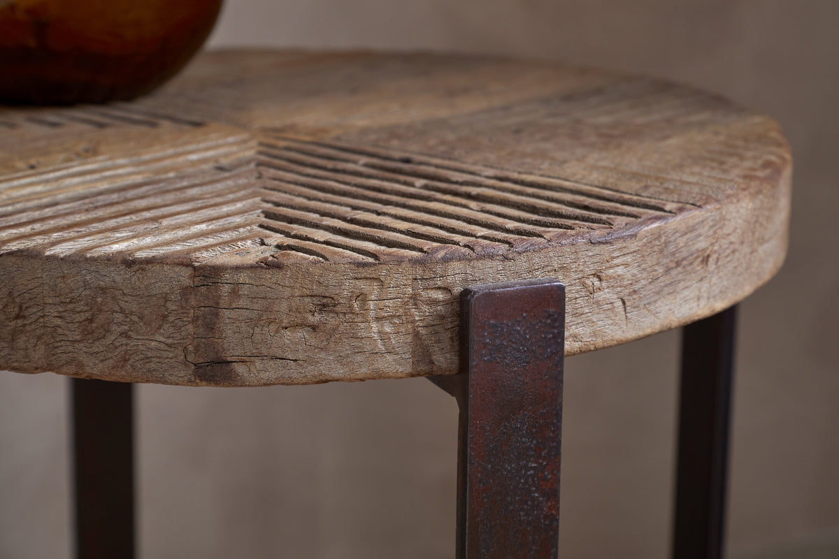 Close-up of the nkuku Chakala Reclaimed Wood Side Table, featuring reclaimed wood with carved geometric grooves, visible grain and cracks, and dark iron legs. Part of a round object is seen on the tabletop.