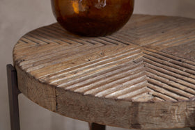 A close-up of the Chakala Reclaimed Wood Side Table by nkuku showcases its carved diagonal grooves and weathered wood. Iron legs and an amber glass vase highlight the sustainable elegance of this reclaimed wood piece.