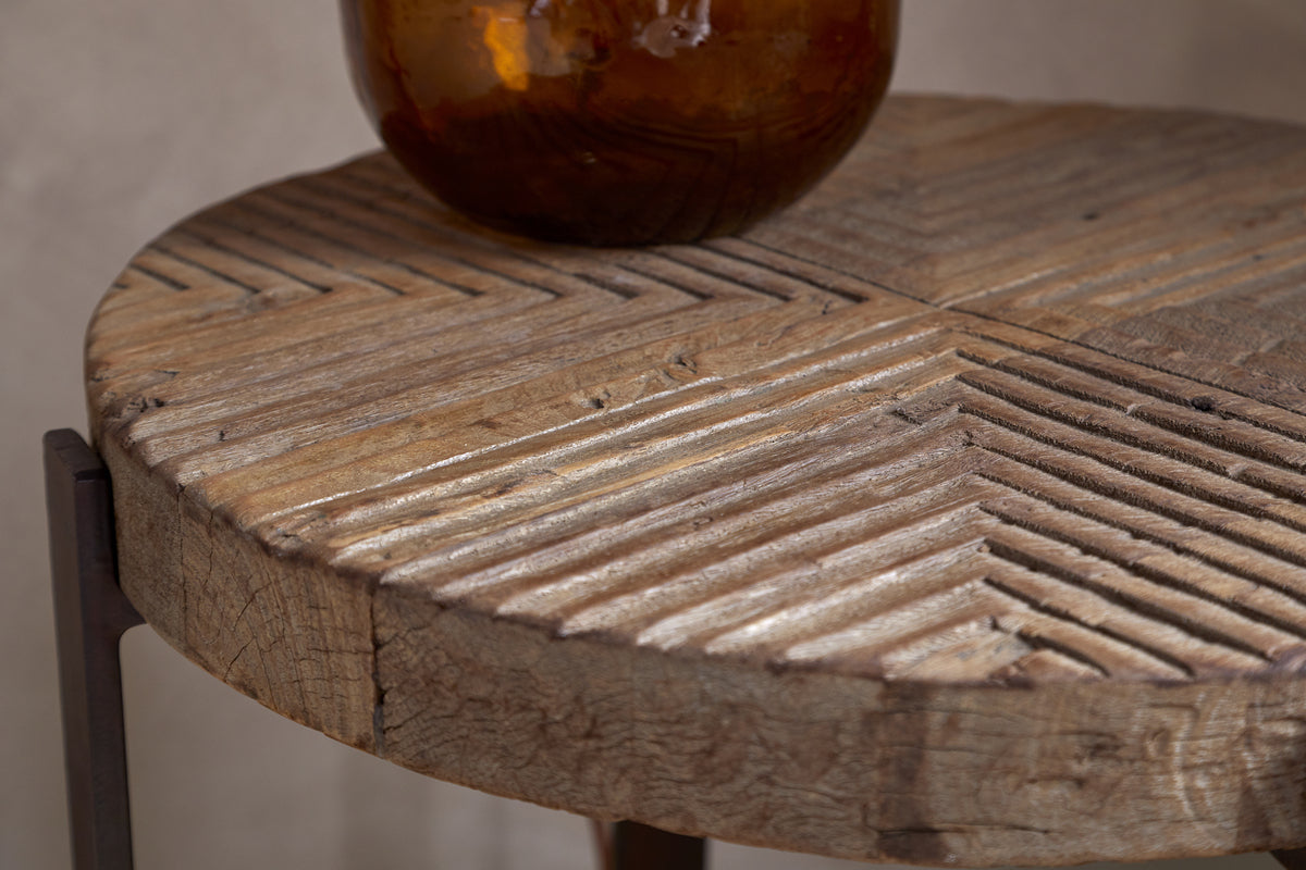 A close-up of the Chakala Reclaimed Wood Side Table by nkuku showcases its carved diagonal grooves and weathered wood. Iron legs and an amber glass vase highlight the sustainable elegance of this reclaimed wood piece.