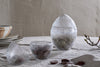 Three nkuku Essan Recycled Glass Opening Egg containers in clear—one open as a lidless egg—hold dark chocolates on neutral linen beside plates and a budding branch, creating a decorative home display.