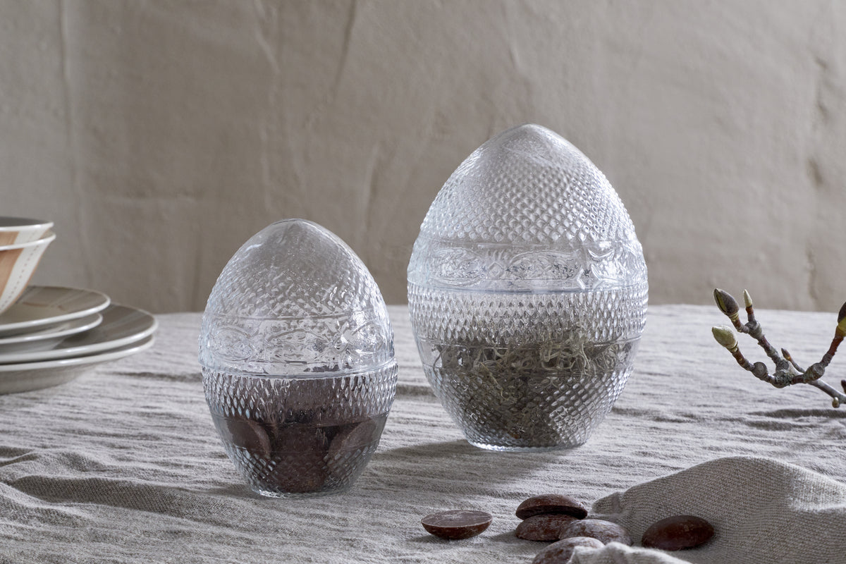 Two Essan Recycled Glass Opening Eggs by nkuku, made from clear textured recycled glass, sit on a light tablecloth. Each jar is filled with brown and green contents—perfect as unique decorative home accents. Plates are stacked left; a twig lies right.