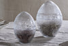 The nkuku Essan Recycled Glass Opening Eggs feature clear, textured recycled glass and decorative bands, making them stylish decor accents for any room. Their unique egg shape stands out beautifully on a beige fabric surface.