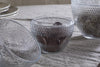 Three Essan Recycled Glass Opening Egg bowls by nkuku sit on a gray linen cloth—one open, another with chocolate and dark filling, the third with dried decor. Stacked plates blur in the background for a decorative touch.