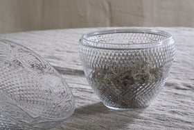 Two Essan Recycled Glass Opening Egg bowls by nkuku, with a diamond-patterned design, rest on a beige textured fabric—one upright holding dried plants, the other tipped on its side. Both are made from clear recycled glass.