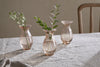 Three nkuku Bapti Bud Vases in Dusty Terracotta sit on a beige tablecloth. Two vases each hold a green sprig—one with drooping white flowers, one with leaves—while the third is empty. A wooden chair appears in the background.