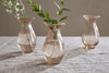 The Bapti Bud Vases - Dusty Terracotta (set of 3) by nkuku sit on a gray textured cloth; two display green leafy stems, the third is empty. Soft natural light creates gentle shadows across the table and background.