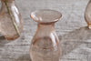 A close-up of an nkuku Bapti Bud Vase in Dusty Terracotta, featuring a translucent light brown tint, wide flared rim, and narrow neck, filled with water. Two similar vases from the set of 3 are partially visible on textured light gray fabric.