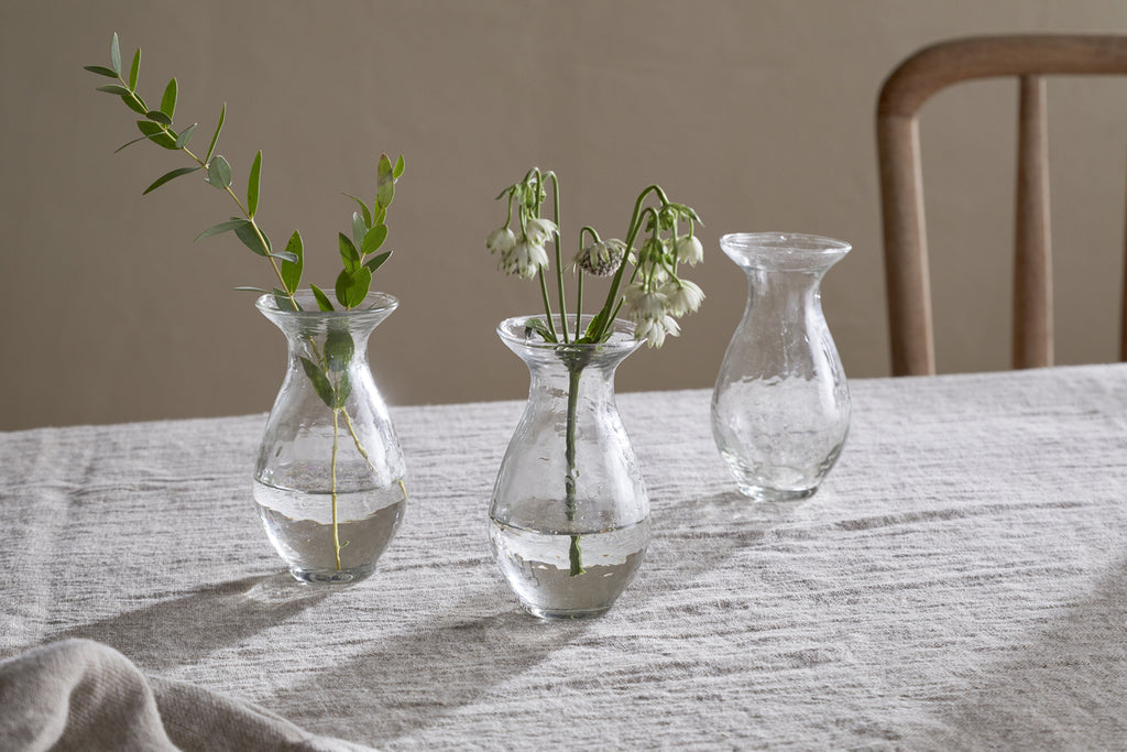 The nkuku Bapti Bud Vases - Clear (Set of 3) are displayed on a light gray linen tablecloth. One holds a leafy sprig, another has delicate white flowers, and the third is empty. A wooden chair can be seen in the background.
