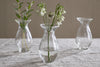 The Bapti Bud Vases (Set of 3) by nkuku sit on a light gray textured tablecloth. The left holds green foliage, the center has white drooping flowers, and the right is empty; all have water, rounded shapes, and flared rims.