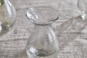 The nkuku Bapti Bud Vases - Clear (Set of 3) feature wide, flared rims and narrow necks. One vase with visible water droplets stands on a light gray textured surface, while two blurred vases are in the background.
