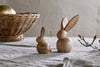 Two nkuku Khara Mango Wood Rabbit Standing Decorations in natural mango wood, one small and one large, are displayed on a textured gray cloth. A wicker basket with a golden accent and budding branches complete the cozy Easter scene.