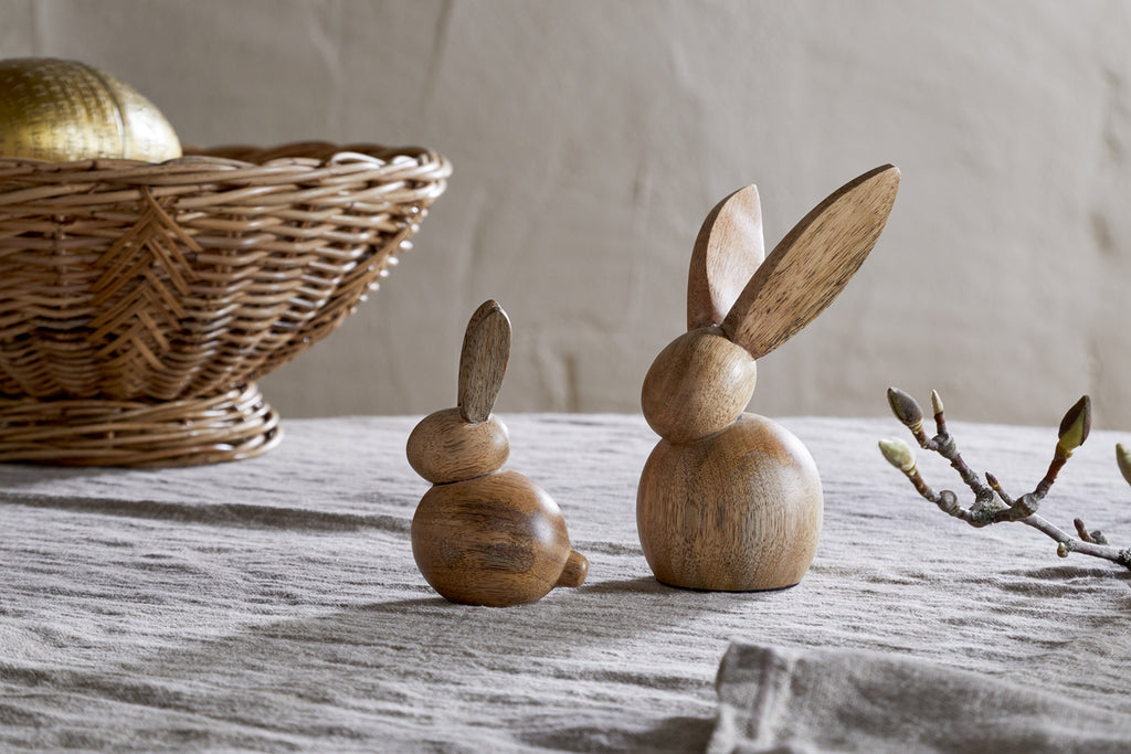 Two nkuku Khara Mango Wood Rabbit Standing Decorations in natural mango wood, one small and one large, are displayed on a textured gray cloth. A wicker basket with a golden accent and budding branches complete the cozy Easter scene.