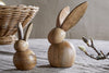 Two Khara Mango Wood Rabbit Standing Decorations by nkuku, featuring smooth rounded shapes and long ears, sit on a grey fabric surface. A wicker basket appears in the background left, with a budding branch to the right.