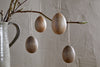 Nkuku’s Dima Mango Wood Hanging Egg Decorations (Set of 4) add minimalist, natural Easter décor when hung from a branch in a gray ceramic pitcher, set against a textured neutral-toned wall.