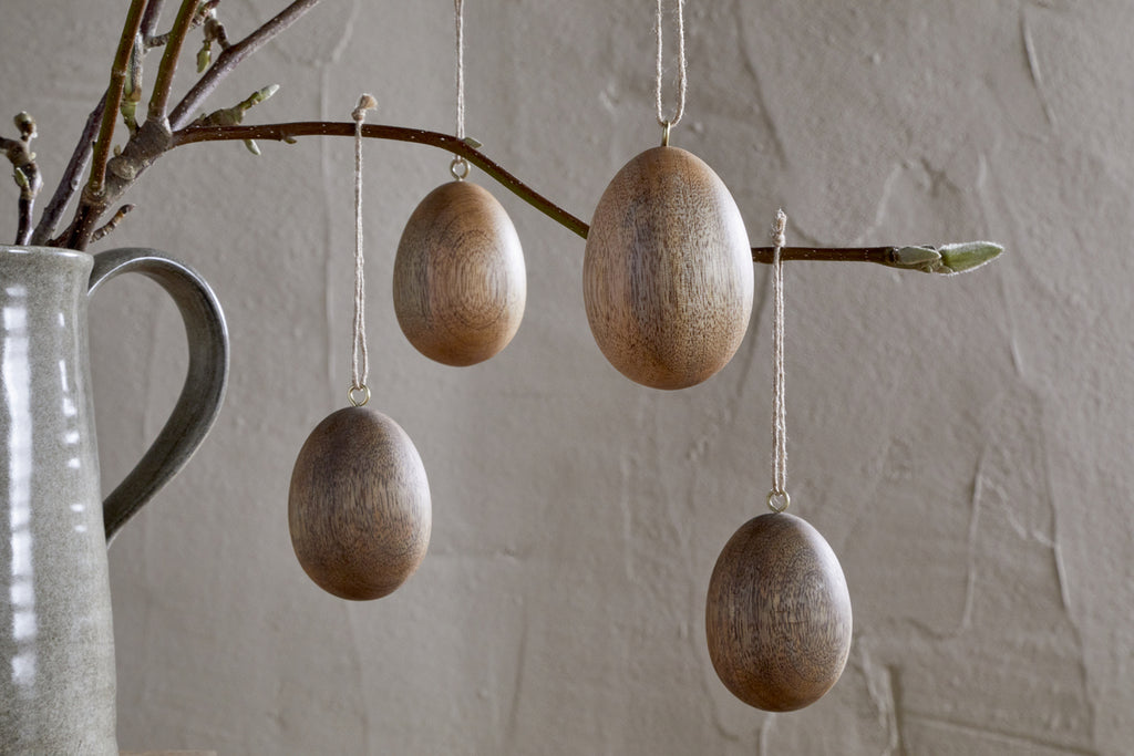 Nkuku’s Dima Mango Wood Hanging Egg Decorations (Set of 4) add minimalist, natural Easter décor when hung from a branch in a gray ceramic pitcher, set against a textured neutral-toned wall.