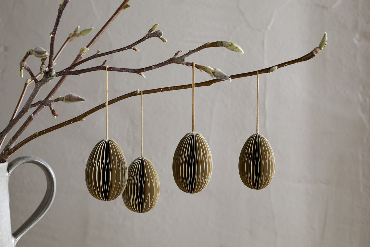 A budding branch in a gray vase displays four nkuku Piani Hanging Paper Decorations in moss green. The recycled ornaments hang on strings, set against a light, textured wall for a minimalist, natural spring look.