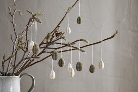 A ceramic jug holds budding branches, decorated with nkuku's Piani Mini Hanging Paper Decoration - Natural & Moss (set of 12), honeycomb eggs, and neutral accents, all set against a light wall for a calm, minimalist scene.