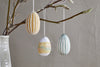 The Terra Hand Painted Recycled Cotton Paper Hanging Egg Set of 3 by nkuku, in teal, cream, and ochre, adds a charming touch to Easter decor when displayed on a branch in a simple white vase against a light wall.