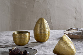 Three Mutti Hammered Opening Eggs in antique brass by nkuku, with intricate engraving, sit on a textured light gray tablecloth. One is open to reveal its interior. A plate of dark treats and a beige bowl with gray napkin are nearby.