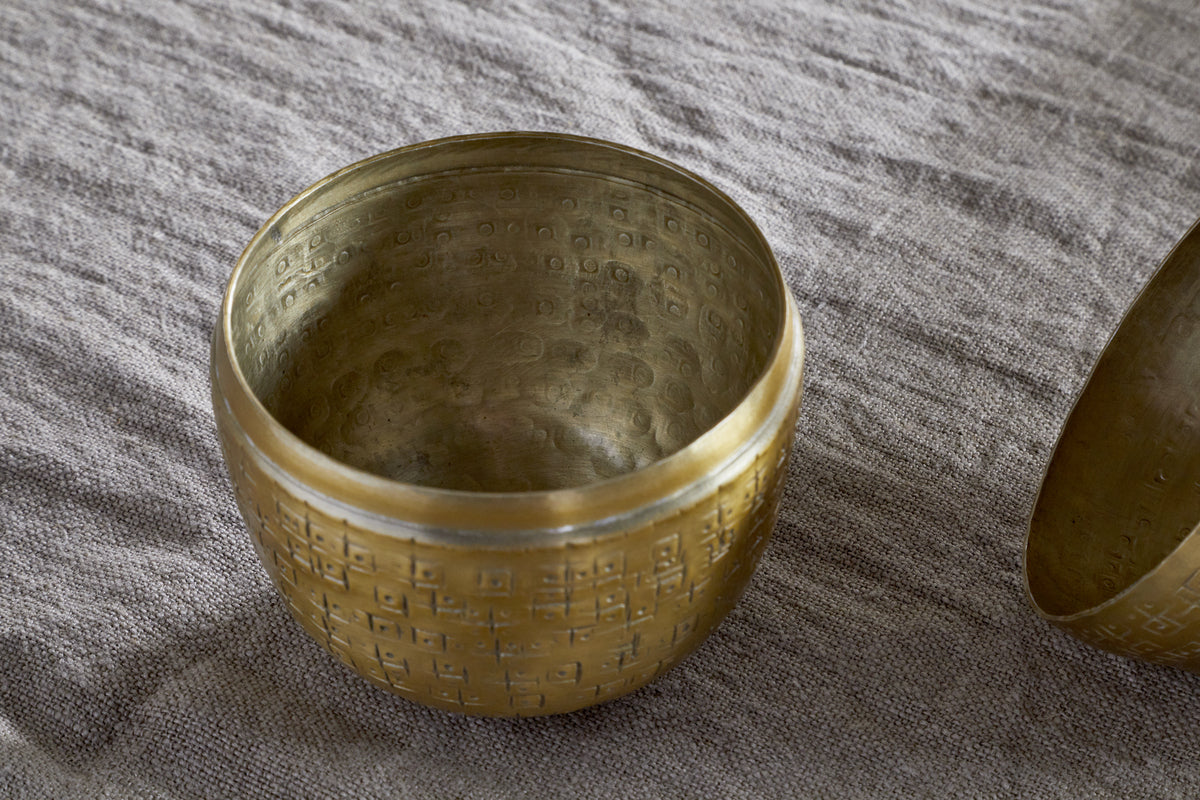 The Mutti Hammered Opening Egg by nkuku, crafted from antique brass with intricate engravings and a softly aged finish, rests on textured gray fabric. A glimpse of a second similar piece appears at the edge, making it a unique seasonal keepsake.
